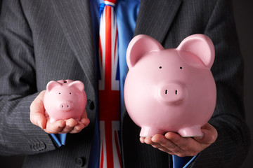 Businessman With Union Jack Tie Holding Large And Small Piggy Bank