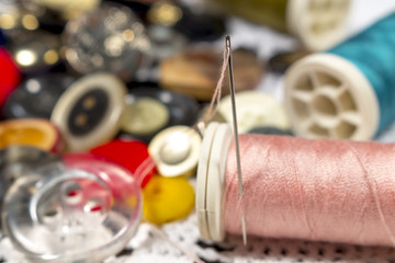 Detail of threaded needle in a spool of pink thread beside different buttons, on a white crocheted...