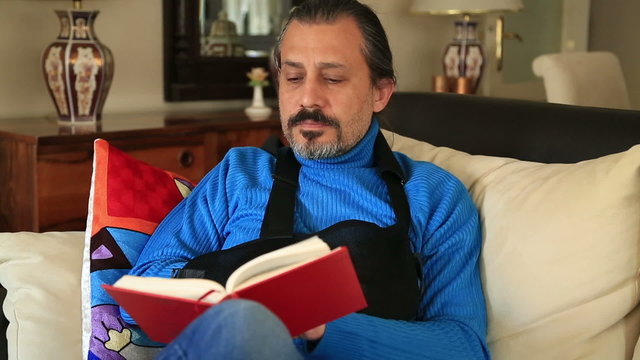 Painful man with injured arm and bandage sitting on sofa and reading a book