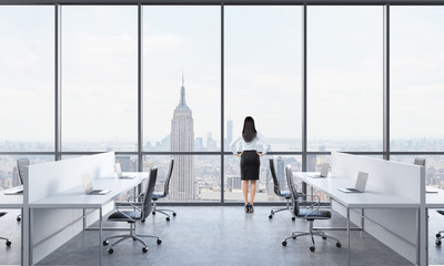 Rear view of a brunette who is looking out the window in the modern panoramic office with New York view. White tables equipped with modern laptops and black chairs. © ImageFlow