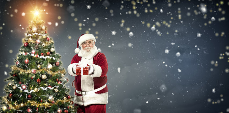 Santa Claus holding gift and standing next Christmas tree