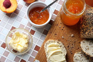 Tasty jam in the jar and bowl, ripe peaches, bread with butter and wooden tablet on mosaic background
