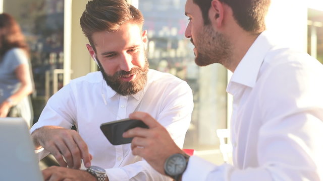 Portrait of two young handsome caucasian business man using smartphone talking to each other in city backlight and tapping the screen – business, work concept – side view