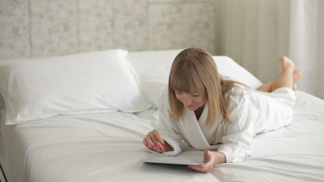 Pretty young woman relaxing on bed using touchpad and smiling