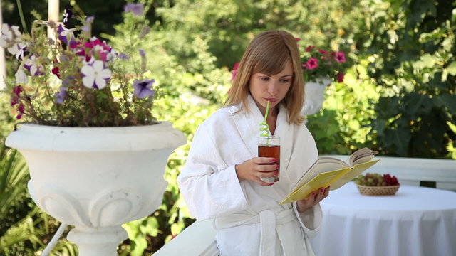 Young woman in a white bathrobe reading a book and drinking apple juice. Young adult standing on the balcony