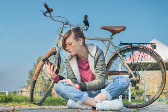 pretty girl reading e-book on a sunny day on clean blue sky with her bicycle in background. eco-conscious modern young woman with sun glasses reading - modern simplicity and clean environment concept 