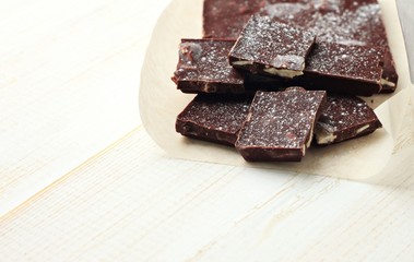 Pieces of black chocolate bar cracked in parchment paper on white wooden table. Copy space.