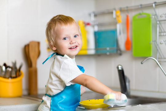 Toddler Child Washing Dishes In Kitchen. Little Boy Having Fun With Helping To His Mother With Housework