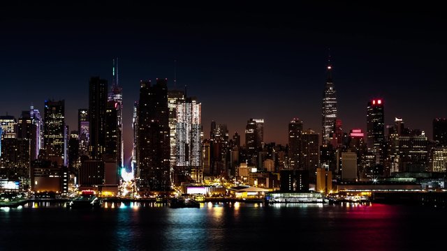 Midtown Manhattan transitions from night to day. A large luxurious cruise ship sails Hudson River waters when sunrise beams shine through the skyscrapers.