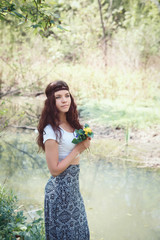 Portrait of  young girl with long dark hair wearing white top, long skirt, hippie headband holding small yellow flowers, standing by river water in forest 