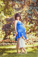 Portrait of caucasian pregnant mother in blue dress in park outside, with daughter in white clothes  on sunny spring summer day, playing, laughing, having fun