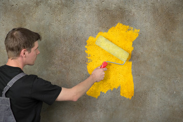 Eine Betonwand wird von einem Arbeiter in grau/schwarz mit der Rolle gelb angestrichen - 96490982