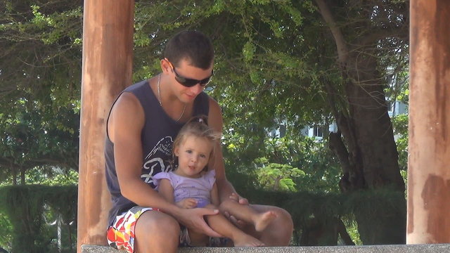 dad and baby playing in the gazebo by the sea. girl stomping feet. Dad keeps daughter's legs. They are laughing.
