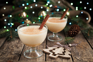 Eggnog traditional xmas homemade winter egg, milk, rum, vanilla alcohol liqueur preparation recipe in two glass cups with cinnamon sticks on wooden vintage table. Blue and green bokeh background