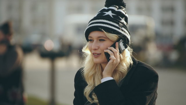 Blonde young woman talking on the mobile phone and smiling while sitting on the edge of parapet