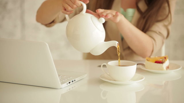 Young woman sitting at table with laptop pouring tea from teapot into cup