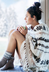Young beautiful brunette woman drinking cup of coffee wearing knitted nordic print poncho sitting home by the window. Blurred winter snow tree background