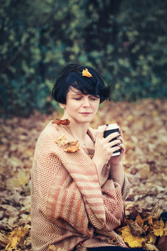 Portrait Of Middle Aged Caucasian Brunette Woman With Leaves In Her Short Bob Hair, Yellow Woolen Scarf, Sitting On Ground Outside In Park Drinking Coffee, Her Eyes Closed, Cozy Autumn Fall Mood