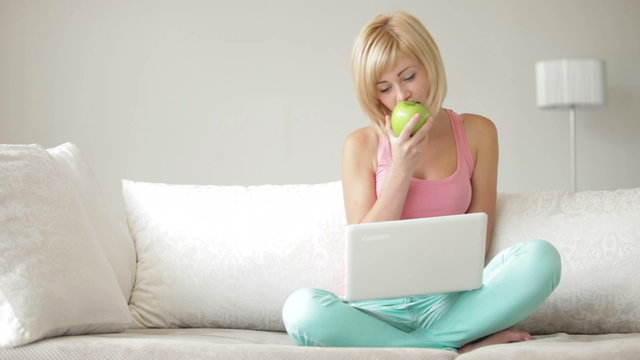 Smiling girl sitting on couch with crossed legs eating apple and using laptop
