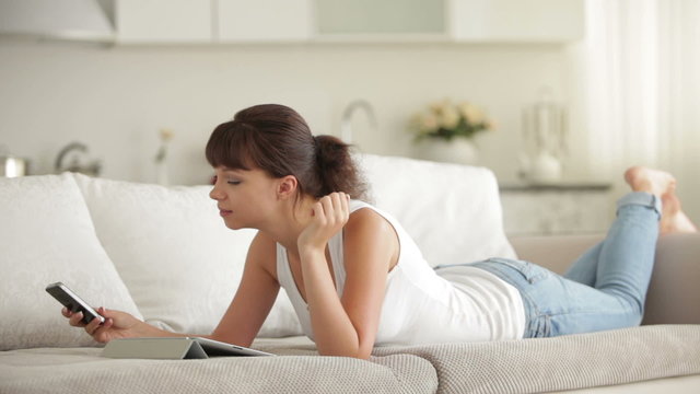 Young woman lying on sofa using touchpad talking on cellphone and smiling at camera