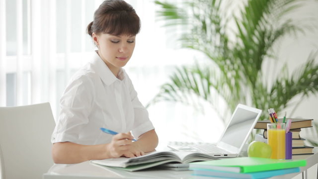 Young woman sitting at table writing in notebook and using laptop