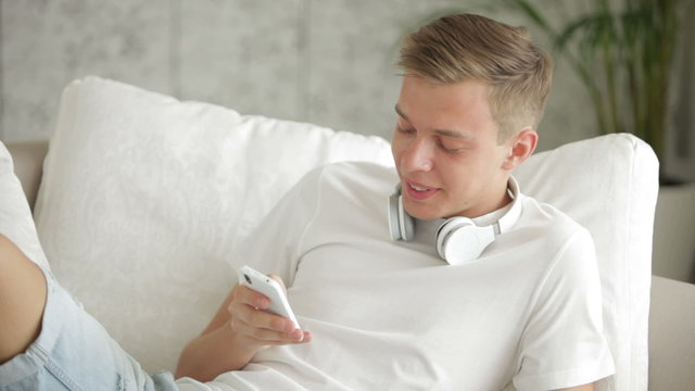 Attractive guy relaxing on sofa using cellphone and eating apple