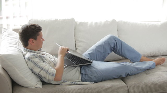 Attractive guy relaxing on sofa using laptop closing it and looking at camera
