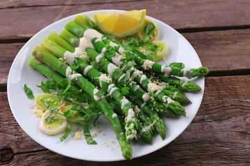 Appetizing asparagus in sour cream sauce with onion, cheese and lemon on white plate against wooden background