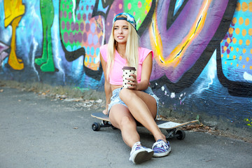 Young woman with skating board and coffee sitting on asphalt on painted wall background