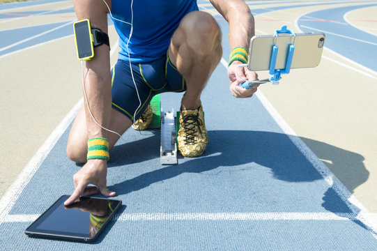 Modern hi-tech athlete crouching at the starting line of a running track wearing a fitness armband, multitasking between using a touchscreen tablet, and taking a selfie with his mobile smartphone 