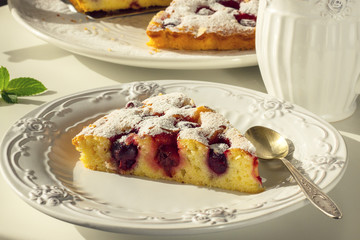 Tea in a white mug, a piece of cherry pie on a white plate on a white table in a range of natural daylight. Horizontal photo.