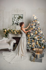 A young girl in a white dress decorates Christmas tree in the living room