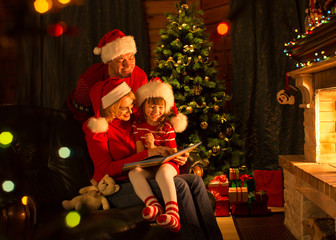 Family reading a book by a Christmas tree in cozy living room in winter