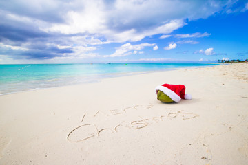 Fototapeta na wymiar Merry Christmas written on beach white sand with red Santa hat