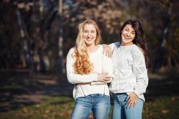 Fototapeta premium Two happy and cheerful young girl students, blond and brunette in coat and jeans laughing in sunny autumn park full of fallen leaves, horizontal pictures