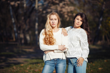 Fototapeta premium Two happy and cheerful young girl students, blond and brunette in coat and jeans laughing in sunny autumn park full of fallen leaves, horizontal pictures