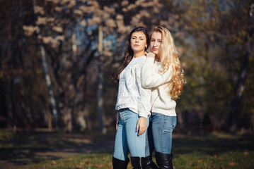 Two happy and cheerful young girl students, blond and brunette in coat and jeans laughing in sunny autumn park full of fallen leaves, horizontal pictures