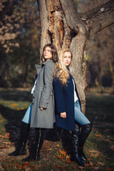 Two happy and cheerful young girl students, blond and brunette in coat and jeans laughing in sunny autumn park full of fallen leaves, horizontal pictures