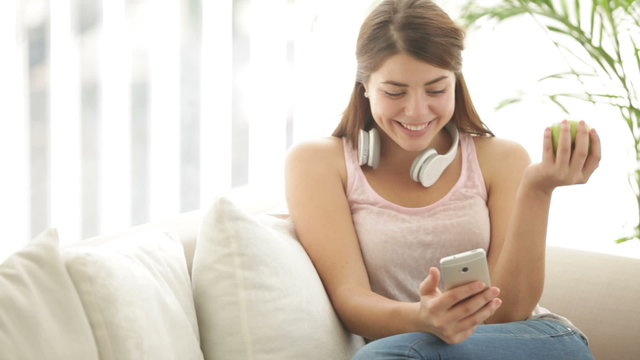 Charming girl wearing headset sitting on sofa eating apple using mobile phone looking at camera and smiling. Panning camera