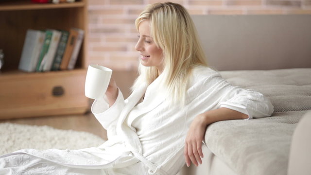 Cute young woman in bathrobe sitting on floor drinking from cup looking at camera and smiling. Panning camera