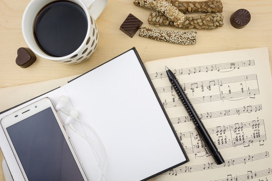 Opened blank notebook with smartphone, pen, cup of coffee and music notation book, on wooden desktop.