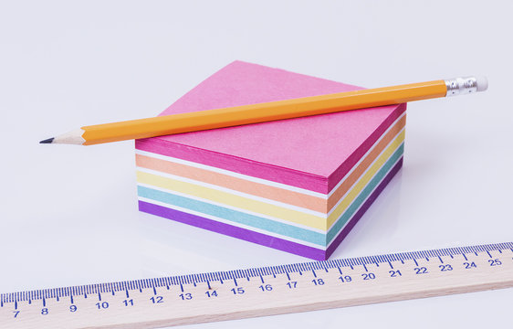 Orange pencil lying on a pack of colored paper on a white table and a wooden ruler