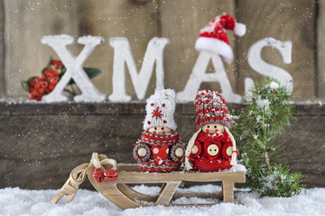 Weihnachtswichtel auf Schlitten im Schnee vor Holzwand mit weißen Holzbuchstaben XMAS