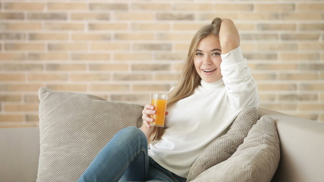 Cute girl sitting on sofa drinking juice looking at camera and smiling. Panning camera