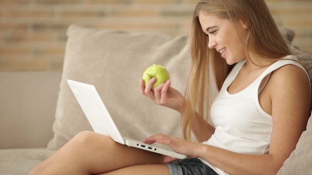 Pretty young woman sitting on sofa using laptop eating apple and smiling. Panning camera