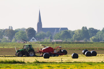 Fototapeta na wymiar Agriculture in the Netherlands with a tractor mowing the field and a church in the background