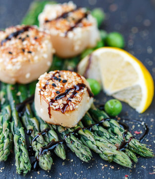Fried scallop  with sesame seeds and balsamic sauce, asparagus, lemon and green peas on a black plate
