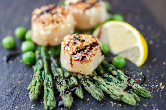 Fried scallop  with sesame seeds and balsamic sauce, asparagus, lemon and green peas on a black plate
