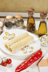 Two crepes with savory filling served on a white plate with marinated mushrooms, pickled cucumber, sour cream and mayonnaise, placed on a white table with spices, vegetables and edible oil.
