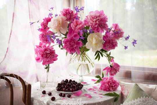 Fototapeta   A bouquet of pink peonies, bells in a vase and cherries in a plate on the table by the window. Still life in a country house. 